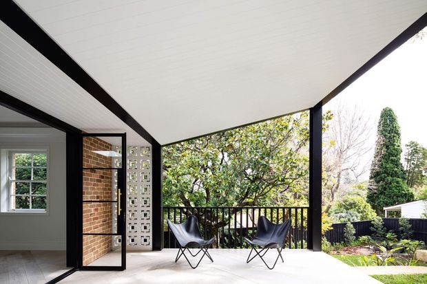 A bright covered outdoor terrace or balcony with a white-lined ceiling, dark structural beams and posts, black balustrade, and pale flooring. Two black sling lounge chairs face the garden. An open glazed door connects the terrace to an interior space with a brick wall and decorative breeze-block screen. Lush landscaped gardens and neighbouring rooflines are visible beyond.
