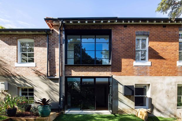 Exterior view of a two-storey brick residential building with a contemporary glazed central extension or entry bay. The facade combines exposed red brick, rendered lower walls, black-framed windows and doors, and a low-pitched roof with visible eaves and timber detailing. A small planted garden and lawn sit in front of the entrance.