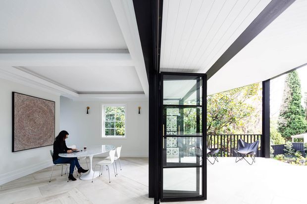 Bright contemporary dining area opening onto a covered outdoor terrace through large black-framed glazed doors. The interior has pale walls, a white dining table with modern chairs, light timber flooring, restrained wall sconces, and a large abstract artwork. The terrace includes black-framed glazing, timber-look decking, railing, and occasional lounge chairs, with greenery visible beyond.