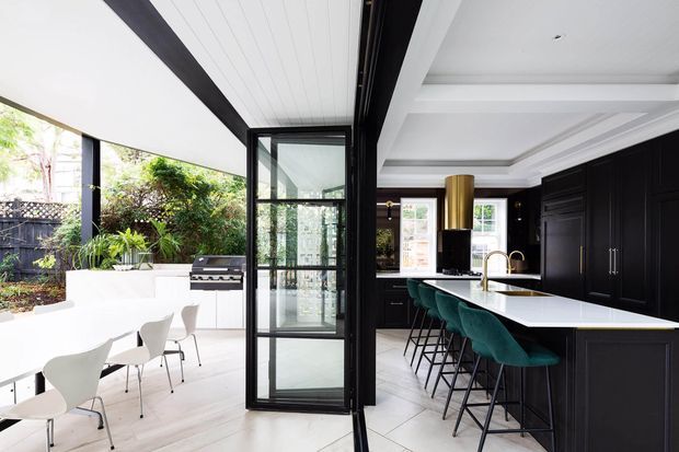 A bright, open-plan kitchen and outdoor entertaining area connected by large black-framed glazed doors. The kitchen features extensive matte-black panelled cabinetry, a long white island with a pale benchtop, teal upholstered bar stools, brass-toned tapware and pendant lighting, and a coffered white ceiling. Outside is a covered dining terrace with white furnishings, built-in planting, and a barbecue. The design has strong indoor-outdoor connectivity and a refined contemporary look.