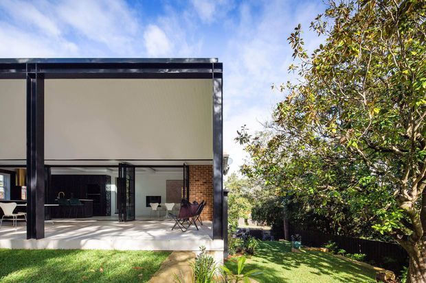 Exterior view of a contemporary single-storey house with a flat roof, dark structural framing, exposed red-brick walls, and large sliding glass openings connecting an open living area to a covered patio. A landscaped lawn and mature tree occupy the foreground and side of the property.
