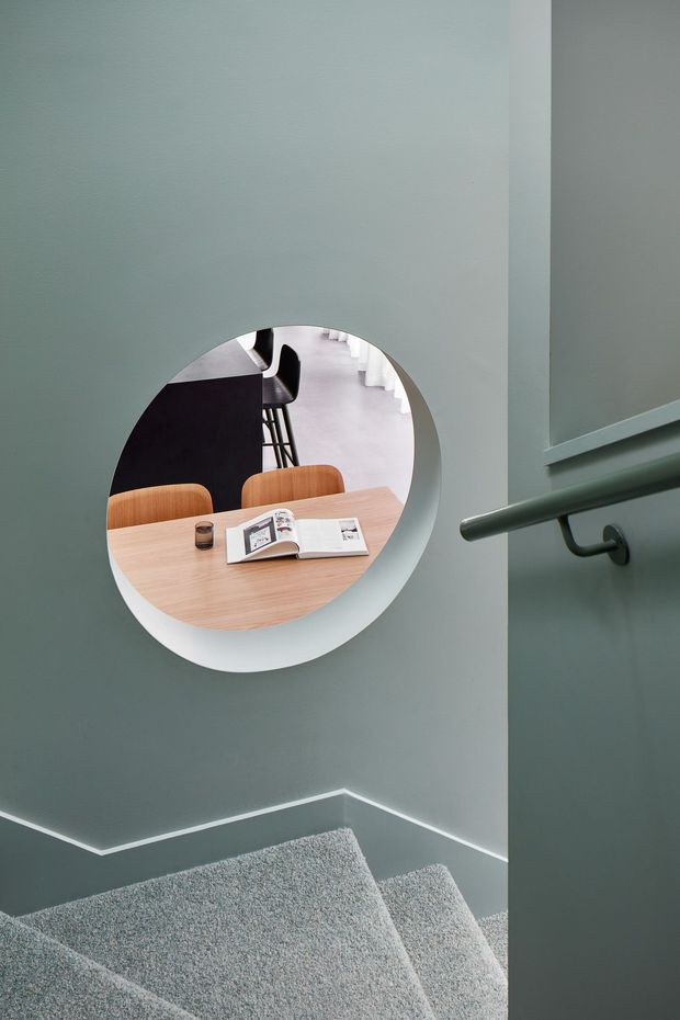 A contemporary stairway viewed from a landing, with muted blue-grey walls, grey carpeted steps, a dark metal handrail, and crisp white skirting. A large circular wall opening frames a view into a dining area with a pale timber table, curved timber chairs, and a dark kitchen island. The composition highlights the playful round aperture and connection between circulation and living spaces.