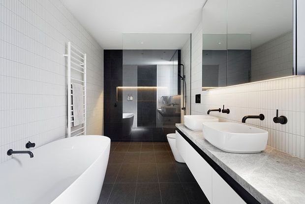 Contemporary bathroom with a freestanding white bathtub, twin vessel basins on a long stone vanity, wall-mounted matte black fixtures, white vertical grid tiles, dark tiled flooring, and a glass-enclosed shower area with dark wall finishes. A large mirror and integrated linear lighting add brightness and depth.