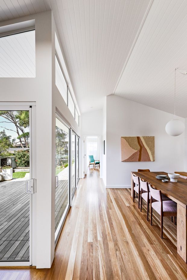 Bright open-plan dining and circulation area with extensive floor-to-ceiling glazing opening onto a timber deck and garden. The room features white vertical-panel ceilings and walls, warm timber flooring, a large timber dining table with upholstered chairs, a sculptural white pendant light, and abstract wall art. A secondary sitting area is visible down the hallway.