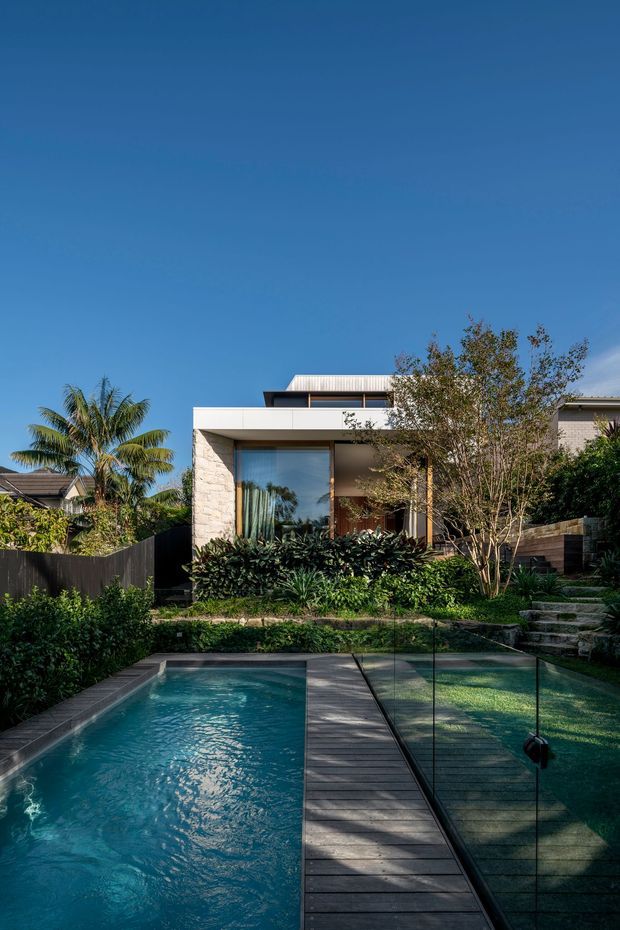 A contemporary residential rear exterior with a rectangular swimming pool in the foreground, crossed by a narrow timber-look deck or bridge and enclosed by clear glass pool fencing. The house has a clean flat-roofed form, pale masonry cladding, large glazed openings, and a landscaped garden with tropical planting and stepped stone paths under a clear blue sky.