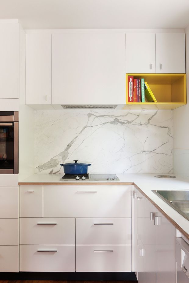 Compact contemporary kitchen with extensive white flat-panel cabinetry, integrated appliances, a white marble-look veined splashback, pale benchtops, stainless-steel sink, and a bright yellow open display niche containing books. A cooktop with a blue pot sits beneath a slim rangehood, and the cabinetry forms an L-shaped work area.