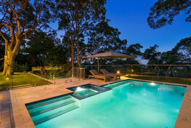 Dusk view of a landscaped outdoor swimming pool area with a large turquoise pool, integrated shallow spa or wading section, broad entry steps, pale stone paving, glass balustrades, sun loungers, and a cantilevered shade umbrella. Mature trees surround the property, with warm landscape and pool lighting creating a relaxed resort-like setting.