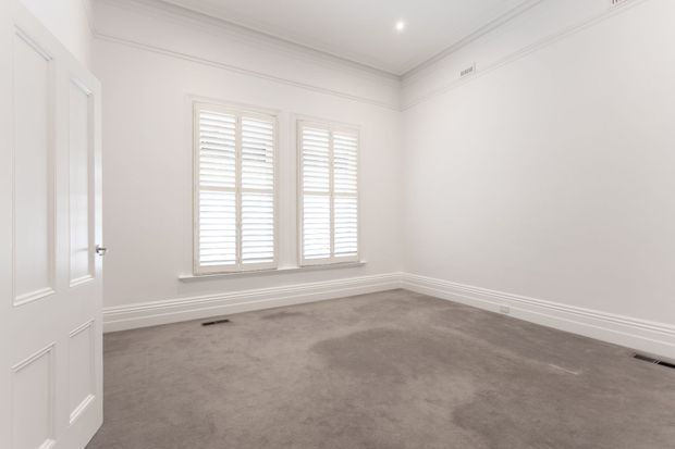 Bright, empty traditional-style room with white painted walls, decorative ceiling cornices and skirting boards, plantation shutters over two windows, a paneled white door, and soft grey carpet flooring.