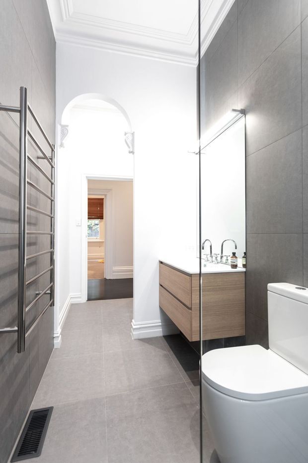 A bright, narrow contemporary bathroom with large grey wall and floor tiles, a wall-mounted timber vanity with white basin and twin chrome taps, a white toilet, frameless glass shower screen, and a tall chrome heated towel rail. White decorative cornices, mouldings, and an arched opening add traditional architectural detailing. A hallway or adjoining room is visible through the doorway.
