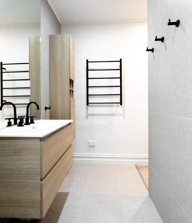 Bright contemporary bathroom with a timber vanity, white benchtop and black tapware, a large wall mirror, pale tiled floor and wall, and black ladder-style wall-mounted towel rails. A tall timber storage cabinet is positioned beside the vanity.