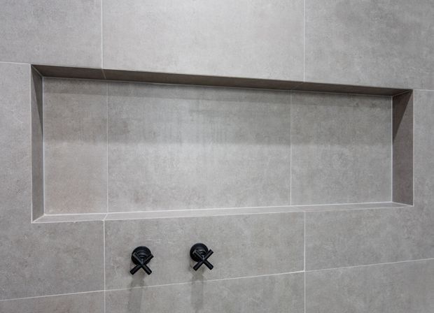 Close-up of a minimalist tiled bathroom wall featuring a long recessed rectangular niche with angled edges and two black cross-handle wall-mounted taps below. The wall and niche are finished in large-format mottled grey tiles.