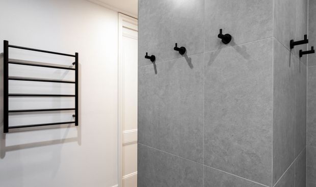 Close-up of a contemporary bathroom featuring large-format grey stone-look wall tiles, matte black wall-mounted fixtures, and a black heated towel rail on a white wall. A partial white paneled door is visible in the background.