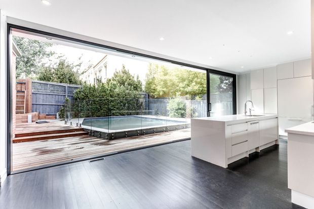Bright contemporary open-plan kitchen with white handleless cabinetry and island, dark timber flooring, and a full-width glazed opening connecting directly to a timber deck and compact swimming pool in a landscaped courtyard.
