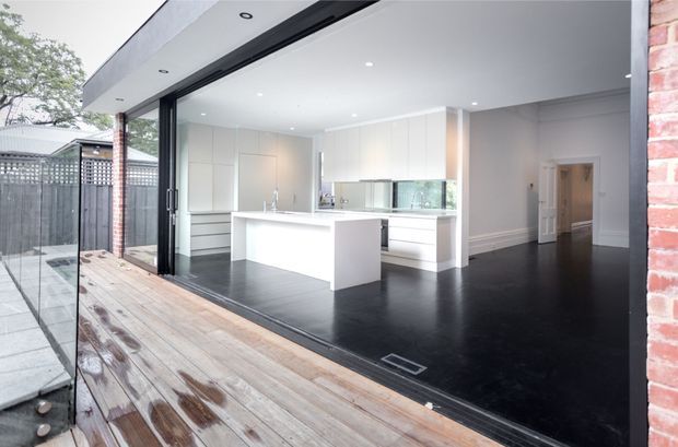 Bright contemporary kitchen opening onto a timber deck through fully retractable glazed doors. The space features white full-height cabinetry, a white island with integrated seating, reflective splashback, dark polished flooring, recessed ceiling lighting, and crisp white wall detailing. A hallway is visible beyond the kitchen.