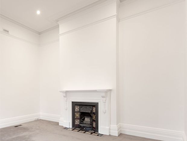 A bright, empty traditional room with white painted walls, ornate ceiling cornices, decorative skirting boards, and a classic mantel surrounding a tiled fireplace. The floor is covered in light neutral carpet, and recessed lighting provides even illumination.