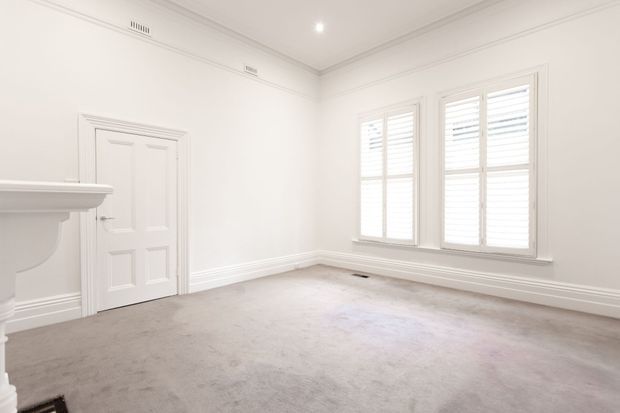 Bright, empty traditional-style room with white painted walls, ornate ceiling cornices and skirting, a decorative white fireplace mantel at left, paneled internal door, plantation shutters, and a soft grey carpeted floor. The room is spacious and receives abundant natural light through two large windows.