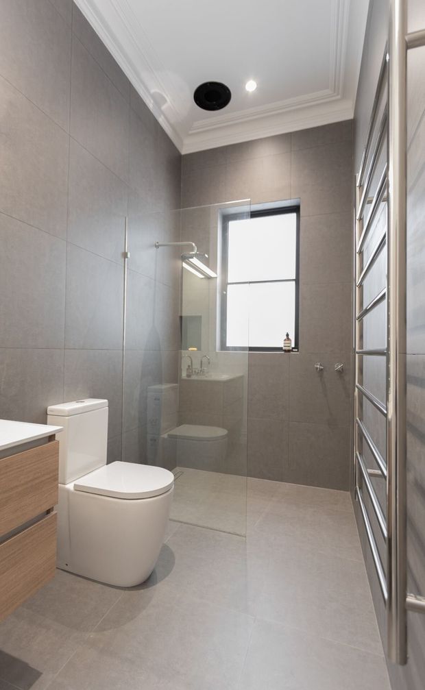 A compact, contemporary bathroom with large grey wall and floor tiles, a wall-hung-style white toilet, timber-toned vanity cabinetry, frameless glass shower screen, chrome tapware, and a tall chrome heated towel rail. The room features a high decorative white cornice ceiling, recessed lighting, and a frosted or bright window.