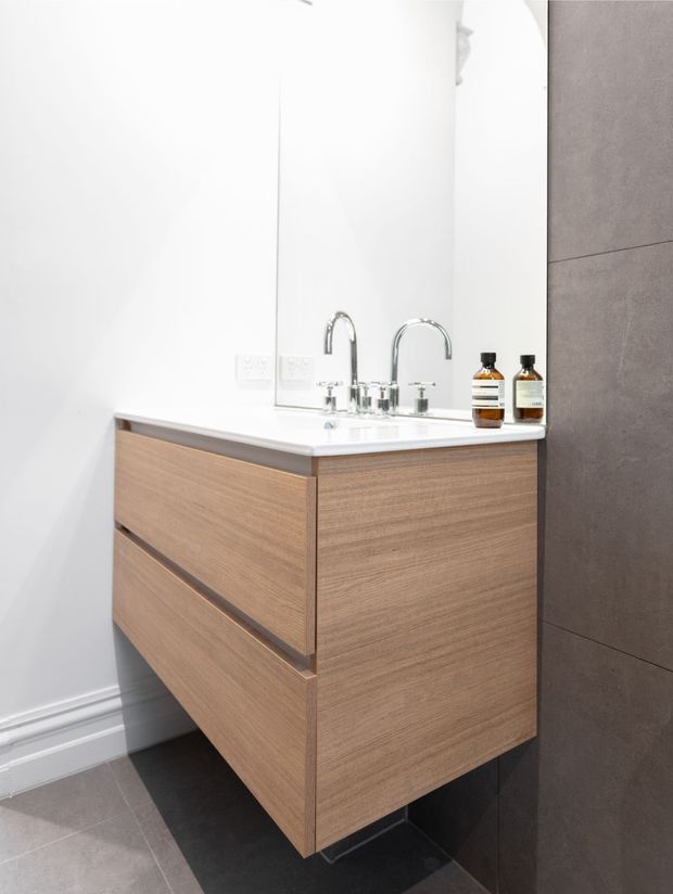 A bright, contemporary bathroom vanity with a wall-mounted light-wood floating cabinet, white integrated basin and benchtop, chrome tapware, and a large mirror. Large dark grey wall tiles contrast with the white walls and timber-look joinery.