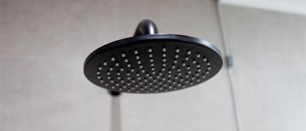 Close-up of a round matte black overhead rainfall showerhead mounted against a light tiled bathroom wall, with part of the shower arm and vertical rail visible.