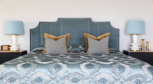 A styled bedroom viewed straight on, featuring a large bed with a decorative blue-grey upholstered headboard, patterned blue and white bedding, textured neutral cushions with mustard trim, and matching bedside tables with lamps. The room has a refined, symmetrical and softly traditional appearance.