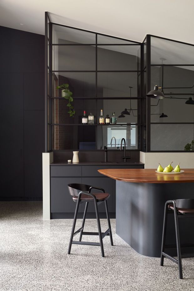 A sophisticated dark-toned contemporary kitchen with matte black cabinetry, a rounded island with a warm timber benchtop, and a black-framed glazed internal partition. The kitchen includes a sink beneath the glazing, open shelving with bottles, black pendant lighting, and two high stools. A speckled terrazzo floor and exposed brick glimpsed through the glazing add texture and contrast.