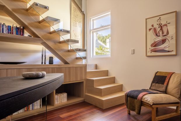 A warm, contemporary interior showing a compact timber staircase with open risers and clear glass balustrades. The stair incorporates built-in timber shelving and cabinetry beneath it, with books and small objects displayed. A round dark dining table occupies the foreground, while a leather lounge chair and framed artwork sit beside a bright window. Rich timber flooring, pale walls, and abundant natural light create a refined, inviting atmosphere.