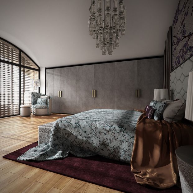 A luxurious bedroom with a large upholstered bed dressed in patterned blue-grey bedding and a brown satin throw. The room features a full-width dark grey concrete-look panelled feature wall with integrated brass wall lights, warm timber parquet flooring, a deep burgundy area rug, patterned accent chairs, and a dramatic crystal chandelier. An arched glazed opening with a perforated screen brings in filtered daylight.