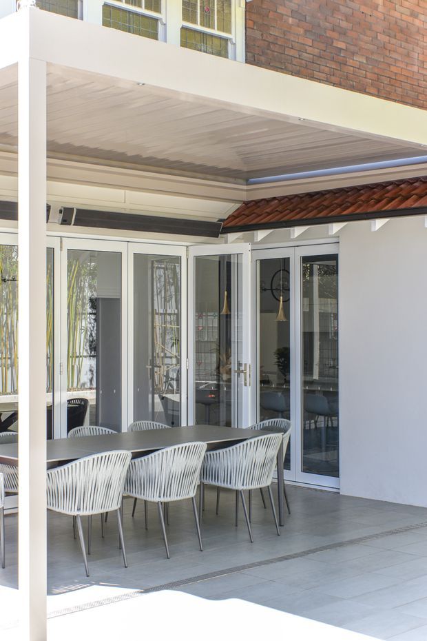 A bright covered outdoor dining area adjoining a glazed interior, with a long dark dining table and white woven chairs on large grey tiled flooring. White-framed folding glass doors open onto the patio, while a red terracotta tiled roof section, exposed white rafters, and brick upper wall add architectural character. The space has a clean, contemporary indoor-outdoor connection.