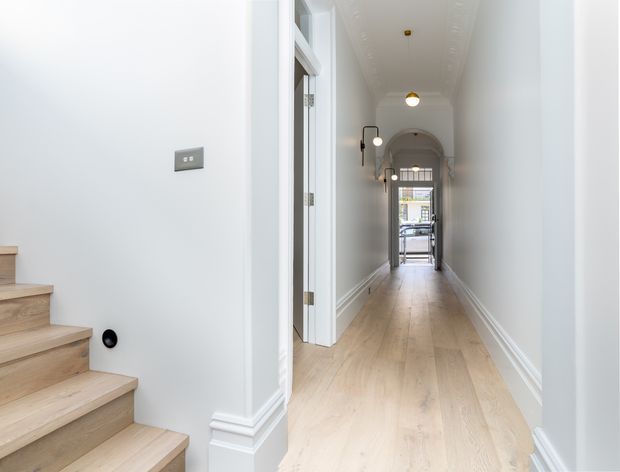A bright, narrow residential hallway viewed from beside a light timber staircase. The corridor has pale painted walls, ornate white skirting and door trim, light timber flooring, recessed wall panelling, arched detailing near the far end, and simple pendant lights. A glazed door or window at the end provides natural light and a view toward the exterior.