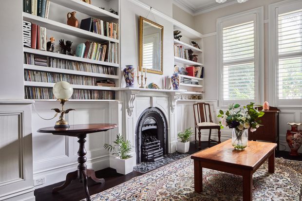 A refined traditional living room with built-in white bookcases filled with books and records, detailed wall panelling, a decorative fireplace with an ornate surround, tall plantation-shuttered windows, and classic dark timber furniture. A patterned Persian-style rug, floral arrangement, ceramics, and framed mirror add warmth and character.
