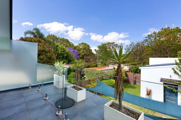 A sunny rooftop terrace or balcony overlooking a landscaped suburban property. The terrace has dark grey tiled or slab flooring, clear glass balustrades with metal posts, and large built-in rectangular planters containing tropical plants. A modern white flat-roofed building with blue accents is visible beyond the terrace, along with lush greenery and a bright blue sky.