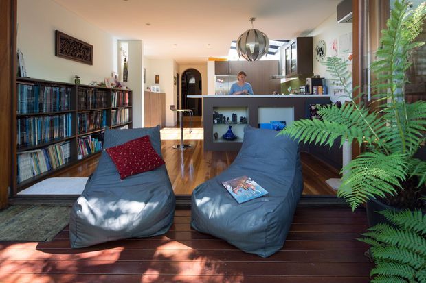 Open-plan living area viewed from a timber-floored glazed sunroom or deck, with two relaxed lounge chairs in the foreground, extensive built-in shelving filled with books and records, and a compact kitchen with island behind. Warm timber flooring, dark cabinetry, large glazing, indoor plants, and a sculptural pendant create a casual, bright, nature-connected interior.