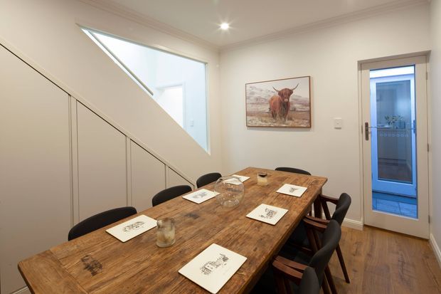 A warm dining room with a long rustic timber dining table and dark upholstered chairs. White walls, timber flooring, recessed ceiling lights, framed artwork, and a glazed door provide a simple, inviting interior. A large angled opening above the stair or adjacent circulation area brings in natural light.