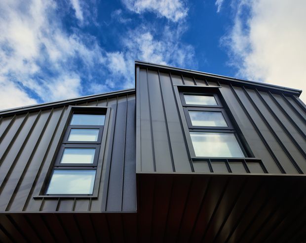 Low-angle architectural photograph of a contemporary multi-level building facade clad in dark standing-seam metal panels. Two vertical groups of dark-framed windows punctuate the angular facade, with a projecting upper volume and metal-lined soffit visible beneath. Bright blue sky and clouds form the background.