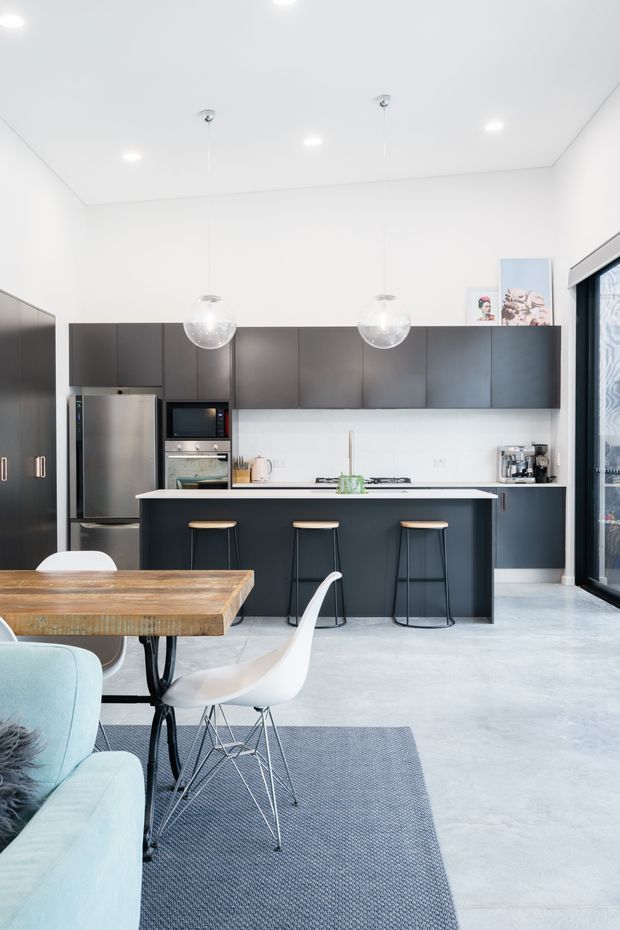 A bright, open-plan contemporary kitchen and dining area. The kitchen features matte dark cabinetry, a white benchtop and splashback, stainless-steel appliances, a central island with three stools, and spherical pendant lights. A timber dining table, white dining chairs, pale blue sofa, grey rug, and large glazed doors complete the clean, airy interior.