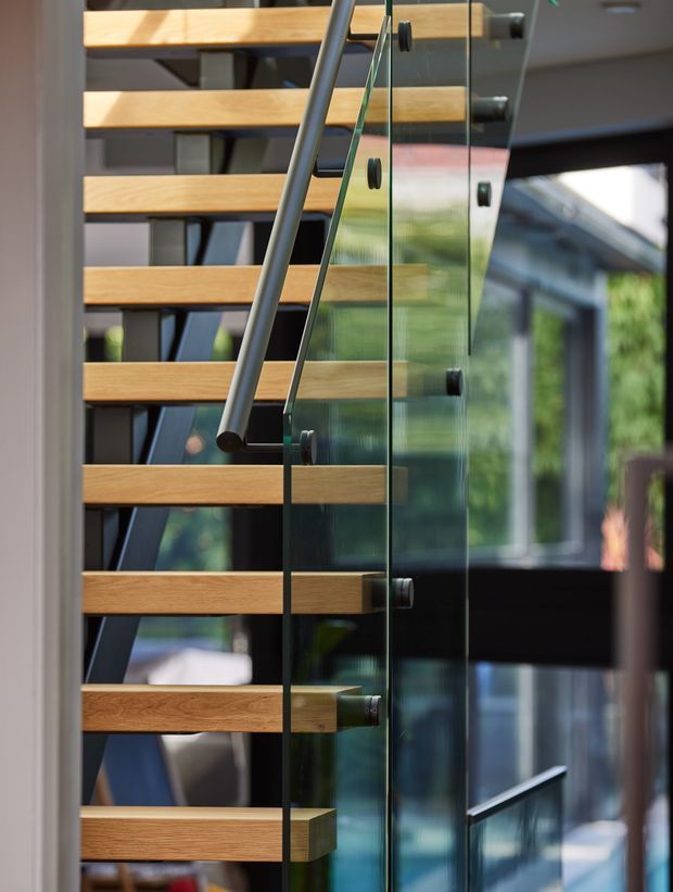 Close-up interior architectural photograph of a modern staircase with open-riser timber treads, dark metal structural supports, and clear glass balustrades secured with dark fittings. The stair is positioned beside large glazed openings, creating reflections and views toward an adjoining exterior or courtyard.