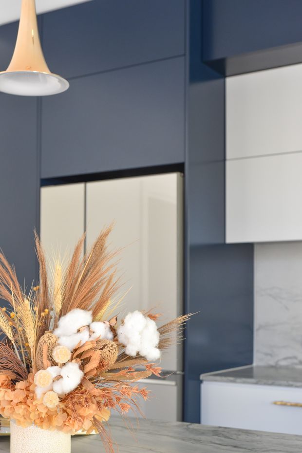 Close-up view of a refined contemporary kitchen with deep navy-blue cabinetry and wall panels, white upper cabinetry, a pale marble-look splashback, and a light grey stone island benchtop. A sculptural pendant light hangs above, while a large dried botanical arrangement in a pale vessel occupies the foreground. The space has a dark, elegant palette balanced by warm natural and cream-toned accents.