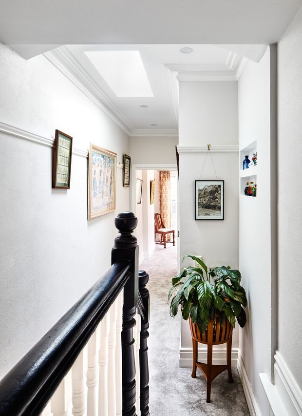 A bright, narrow upper-floor hallway or landing viewed beside a staircase. The space has white textured walls, elaborate white cornices and wall mouldings, recessed ceiling lighting, a large skylight, framed artwork, and a black polished timber stair handrail with turned balusters. A leafy indoor plant in a wooden stand occupies the landing, while a carpeted corridor extends toward a sitting area in the background.