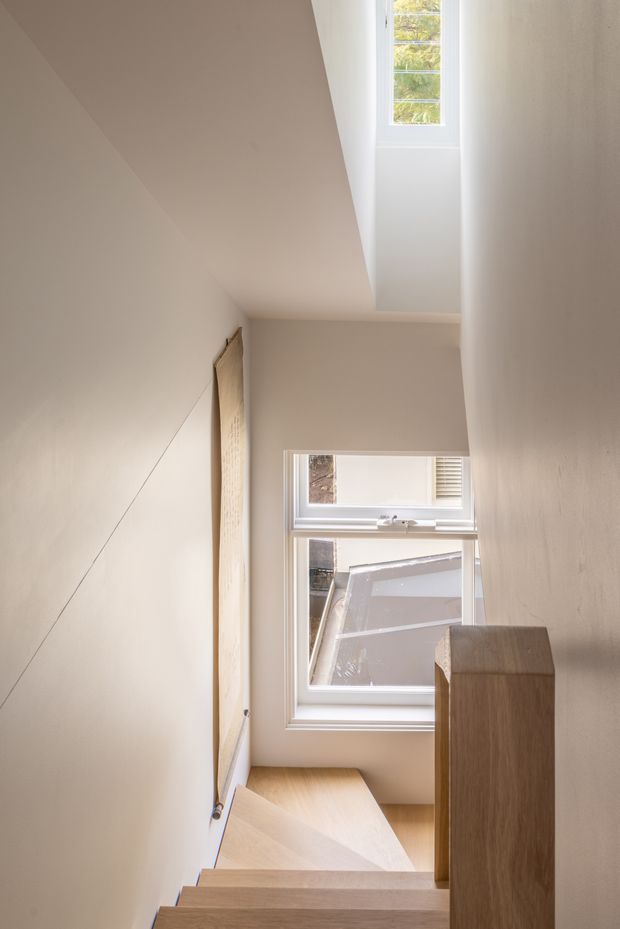 A narrow contemporary stairway viewed from above, with light timber stair treads and a matching timber balustrade or screen. Smooth warm off-white walls and a sloping ceiling frame a high-level skylight and a window overlooking neighbouring rooftops, creating a softly daylit vertical circulation space.