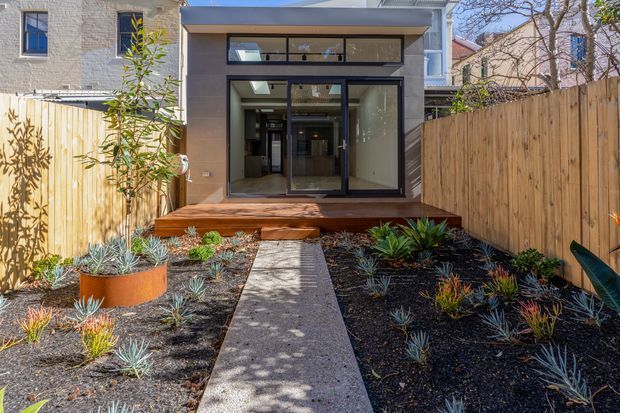 Rear exterior view of a contemporary residential extension opening onto a compact landscaped courtyard. The facade features dark-framed floor-to-ceiling glazing, clerestory windows, a shallow projecting roof, timber decking and steps, and pale rendered walls. A concrete path leads through dark mulch beds with drought-tolerant planting, enclosed by timber privacy fencing.