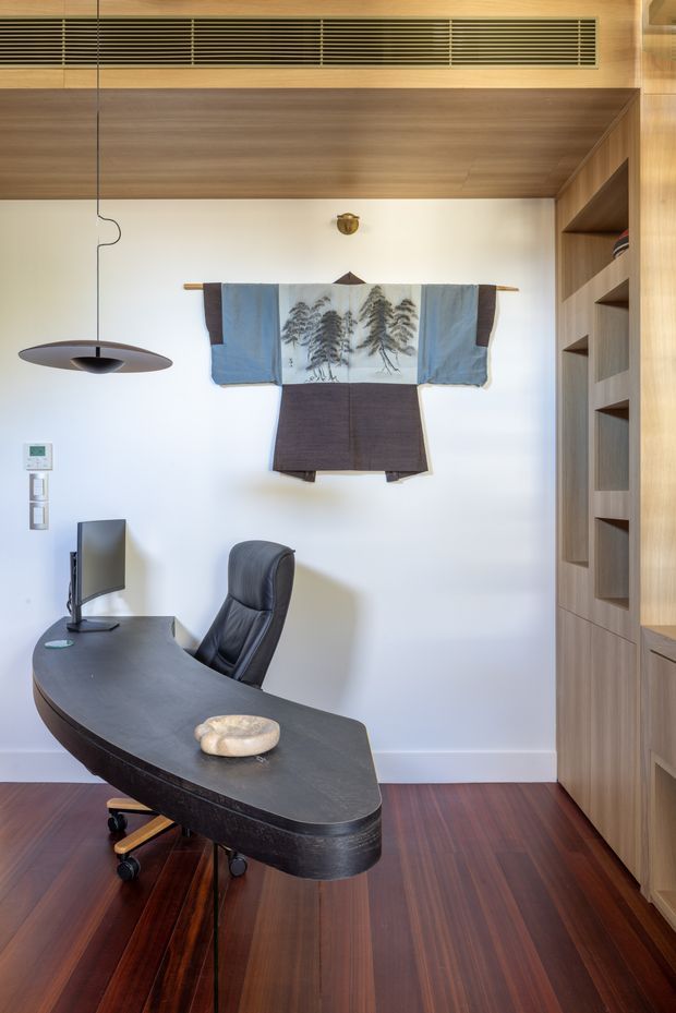 A compact, refined home office with a curved dark desk, ergonomic leather office chair, monitor, and sculptural pendant light. The room features rich dark timber flooring, extensive warm timber built-in shelving and cabinetry, a timber-lined ceiling with a linear ventilation grille, white walls, and a decorative textile wall hanging.
