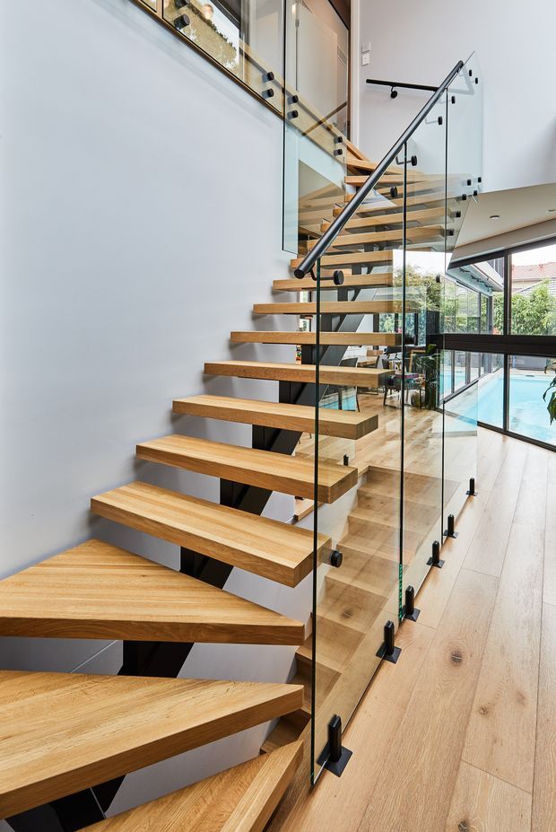 Modern interior stairway with floating solid timber treads supported by a dark structural spine, enclosed by clear glass balustrades with black metal fixings and handrails. The staircase rises through a bright, open double-height space with pale walls and matching timber flooring. Large glazing overlooks a swimming pool and garden beyond.