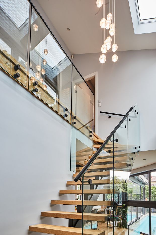 A modern interior stairway with open-riser timber treads, black metal handrails, and clear glass balustrades. The stair rises through a tall, light-filled void with a large cluster pendant light, skylight, extensive glazing, and views toward a pool and garden beyond. A mirrored or reflective glass balustrade visually doubles the space and lighting.