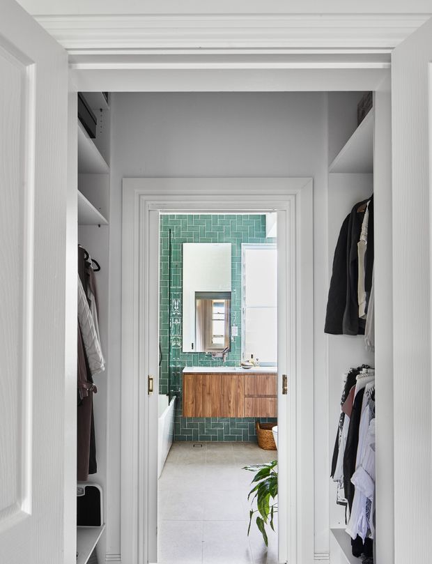 Bright, refined walk-in wardrobe with built-in open shelving and hanging rails, viewed toward an adjoining bathroom. The bathroom features a floating timber vanity, white basin, brass fittings, pale floor tiles, and a striking green vertically arranged glazed-tile wall. White trim, panelled doors, and ceiling detailing create a classic architectural backdrop.
