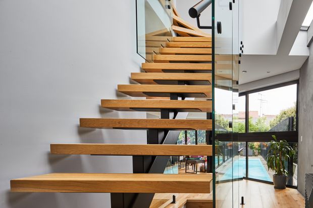 Contemporary interior view of a floating timber staircase with thick open-riser treads supported by a dark structural frame. Clear glass balustrades and large black-framed windows create a bright, connected stairway overlooking a swimming pool and outdoor area. The space uses clean white walls, natural timber, black metal detailing, and abundant glazing.