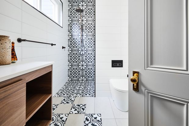 Bright, compact bathroom viewed through an open paneled door. The room features white tiled walls and floor, a floating timber vanity with an open shelf, black tapware and towel rail, a wall-hung toilet, and a striking black-and-white patterned tile feature running vertically behind the toilet and across the floor. A high window provides natural light, while the door has a grey painted finish with brass hardware.