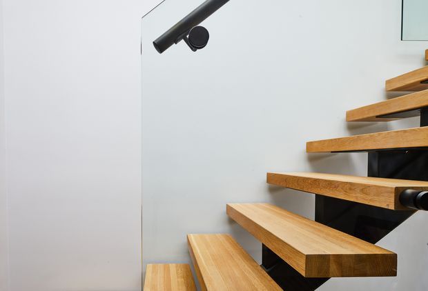 Close-up interior view of a modern floating timber staircase with thick oak-toned treads cantilevered from a dark central structural element. The stairway is set against smooth white walls and includes minimalist black cylindrical lighting and handrail details, creating a clean, contemporary architectural composition.