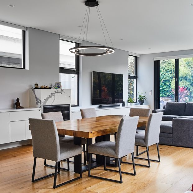 A bright, open-plan dining and living area with a large solid-wood dining table surrounded by upholstered grey dining chairs. White built-in cabinetry, a marble-look fireplace surround, wall-mounted television, grey sectional sofa, expansive windows and glazed doors, and a contemporary circular pendant light create a clean, refined interior.