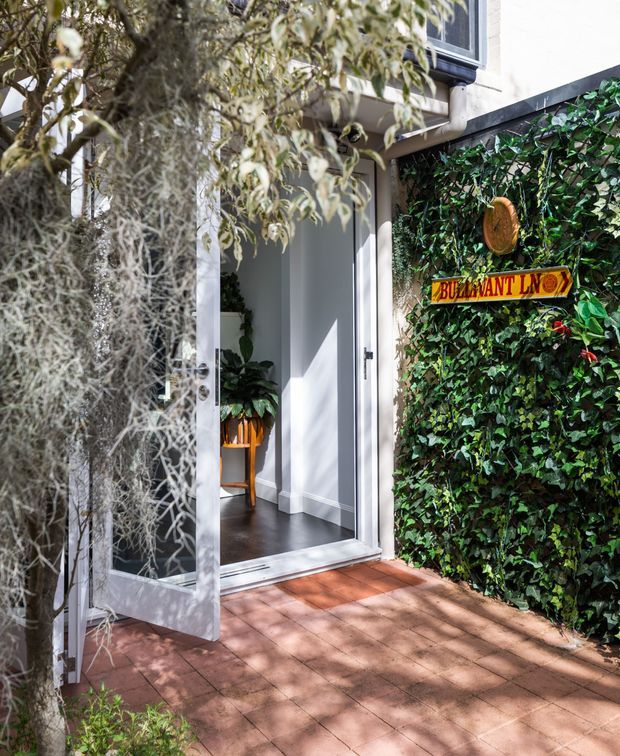 A sunny residential entryway viewed from the exterior, with an open glazed door leading into a compact interior foyer. The entrance has white trim and walls, dark flooring, a small wooden plant stand, and a red-toned brick-paved approach. A dense green vine-covered wall sits beside the doorway and carries a colourful directional sign, while hanging and surrounding foliage frame the scene. The image has a warm, leafy, inviting character.