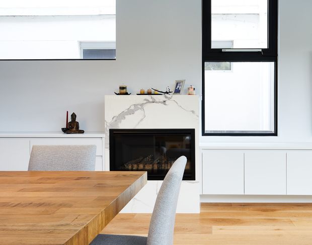 Bright contemporary dining and living area with a large solid timber dining table in the foreground, pale upholstered chairs, white built-in cabinetry, a black-framed window, and a fireplace set into a white marble-look surround. The room has light timber flooring, white walls, and abundant natural light.